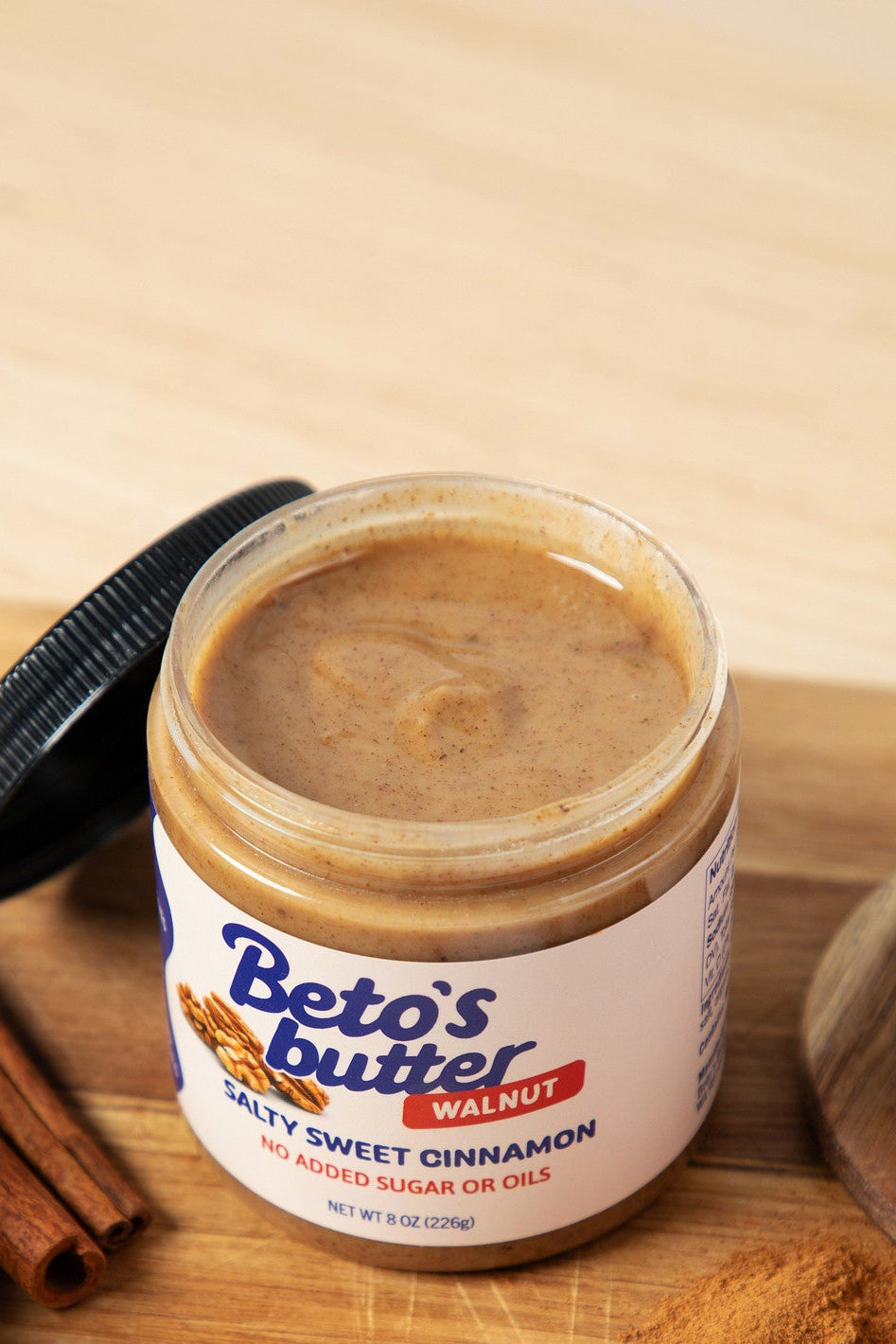 An open jar of Beto's Butter spread on a wooden board, with cinnamon sticks, walnuts, and a small pile of cinnamon powder around it on a light wood surface.
