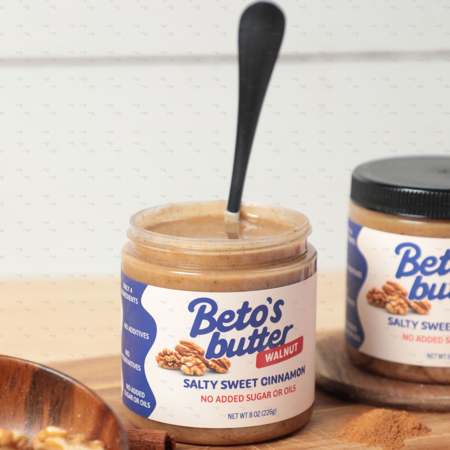 Jar of Beto's Butter with a spoon inside, surrounded by walnuts and cinnamon sticks on a wooden board.
